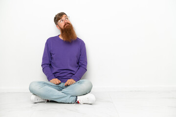 Fototapeta premium Young caucasian reddish man sitting on the floor isolated on white background and looking up