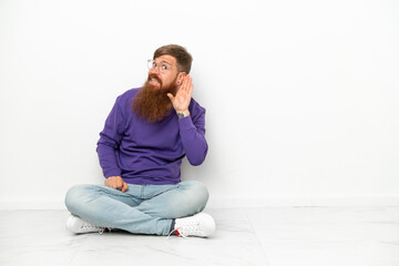 Young caucasian reddish man sitting on the floor isolated on white background listening to something by putting hand on the ear