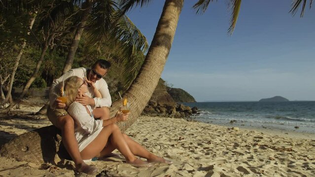 Loving couple in white dress and sunglasses, near palm tree, kissing, relax and drinking cocktail Pina Colada. Concept sea beach resting tropical tourism summer holidays wedding love family vacations