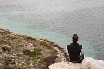 Fototapeta na wymiar Back view of young man sitting on rocks on cliff. Happiness, travel alone and freedom concept.