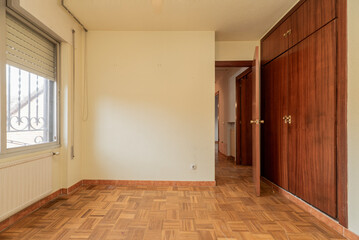 Empty vintage bedroom with aluminum radiator under the window, fitted wardrobe with four wooden doors, oak parquet floor and cream painted walls