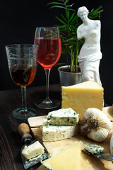 Manchego cheese board combined with blue cheese and various cheese knives, Parmesan cheese shavings, some strong garlic and wine glasses, a potted palm tree and a Greek female figure