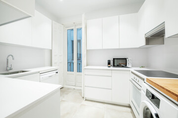Nice kitchen of a short-term rental apartment with white furniture and appliances, with a balcony with vintage wooden doors and a ceramic hob