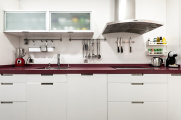 Front image of a kitchen with white lacquered furniture, a red countertop and many accessories on the walls and on the countertop