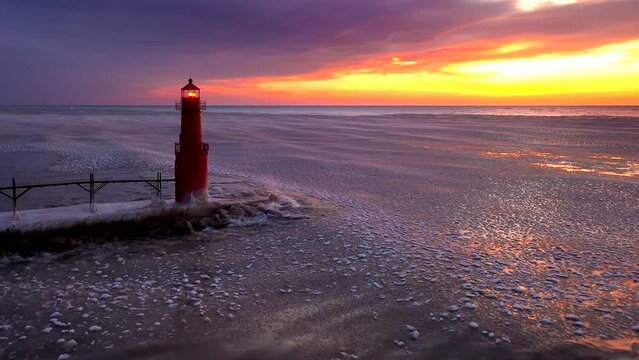 There is amazing beauty in this aerial view of an icy harbor in Winter, clouds on the horizon, waves reflecting the fiery sky, lighthouse beacon blinking, Lake Michigan in the Winter just before sunri