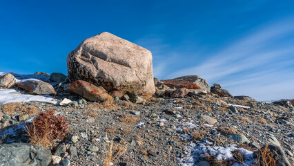 Dry grass, lichens on rocks, snow on the ground are visible on the hillside. A large boulder against a background of blue sky and radiant clouds. Copy space. Altai