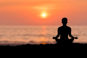 Silhouette of young female sitting practices yoga and meditating in lotus position alone on the rock in the morning at the sea with beautiful sunrise and orange sky. She felt calm and happy.