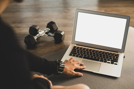 Young sporty woman in black sportswear is sitting on the floor with dumbbells and is using a laptop at the gym after exercise. Sport and recreation concept.