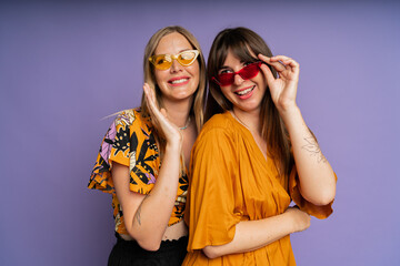 Obraz premium Close up portrait of two stylish women in sunglasses and trendy summer clothes posing on purple bakground in studio.