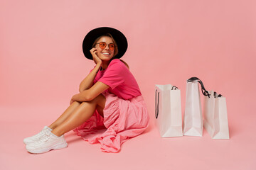 Pretty smiling woman in  spring trendy outfit posing with white shopping bags in studio over pink...