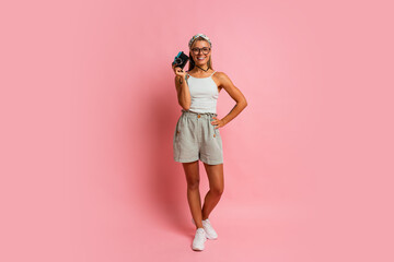 Fototapeta na wymiar Laughing blond woman holding retro camera and posing on pink background. Travel and summer vacation concept. Full lenght.