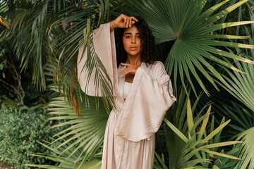 Close up portrait of  blissful  woman with wavy hairs posig  in tropical garden. Wearing elegant...