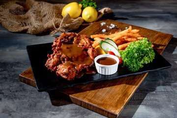 Crispy Boneless Chicken Steak in a black dish with french fries, sauce and lime top view on dark background