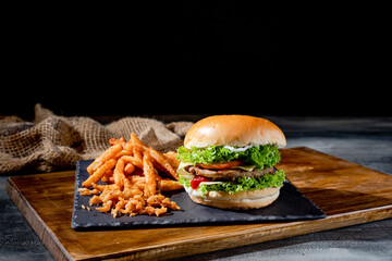 Delicious beef burgers with tomato, cheese, ketchup and french fries on wooden cutting board side view on dark background
