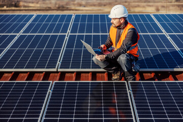 A worker testing solar panels on the laptop while crouching on the roof.
