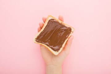 Young adult woman hand holding fresh slice of white bread with brown chocolate cream on light pink table background. Pastel color. Closeup. Sweet snack. Top down view.