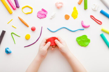 Baby hands holding and kneading red modeling clay on white table background. Closeup. Point of view shot. Toddler development. Making different colorful shapes. Top down view. - obrazy, fototapety, plakaty