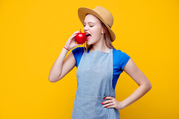 Caucasian female gardener eat apple dressed in straw hat and an apron on yellow background