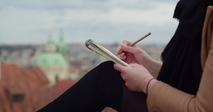 Prague view in background and girl holding paper notepad and pencil, working outside, writing ideas, closeup view