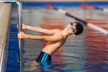A little boy in swimming goggles is holding on to the pool railing.