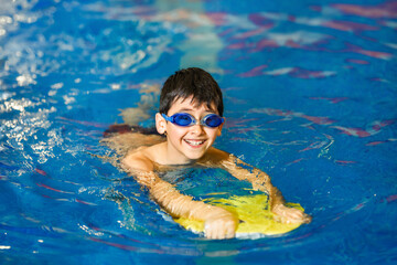 A little boy of 10 years learns to swim in the sport pool using a board. The boy wore swimming goggles. The child is cheerful and happy. Healthy kid enjoying active lifestyle. - Powered by Adobe