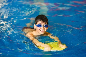 Fotobehang A little boy of 10 years learns to swim in the sport pool using a board. The boy wore swimming goggles. The child is cheerful and happy. Healthy kid enjoying active lifestyle. © Olha