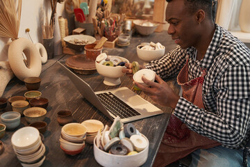 Joyous ceramist seated at laptop showing earthenware products to customer