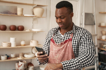 Cheerful potter using his gadget in studio