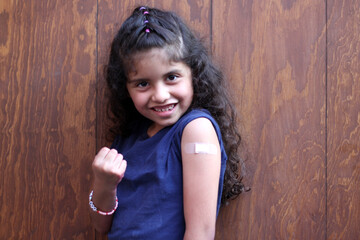 Little latin girl with curly hair happy shows her arm recently vaccinated against Covid-19 in the...