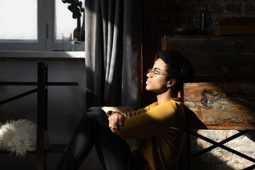 Obraz premium Peaceful happy young African woman, millennial girl sitting on floor in loft interior room, leaning back on shabby wooden furniture, relaxing with closed eye with day light shining on face