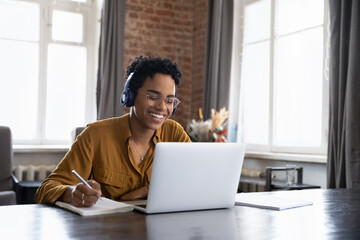 Fototapeta Cheerful Black college student in earphones attending virtual class, writing notes, watching webinar on Internet, studying online. African worker in headphones talking to customer on video call obraz