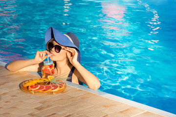 Young girl in a blue hat relaxes in the pool and drinks a cool cocktail of fresh fruit.