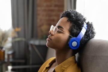 Peaceful relaxed African girl in wireless headphones sleeping to lounge music songs, resting on sofa at home. Calm sleepy fan listening to chill out tunes with closed eyes for relaxation