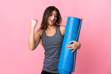 Young sport woman going to yoga classes while holding a mat isolated on pink background celebrating a victory