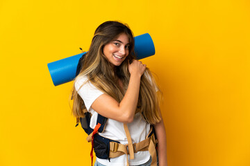 Young mountaineer woman with a big backpack isolated on yellow background laughing