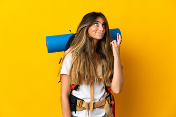 Young mountaineer woman with a big backpack isolated on yellow background with fingers crossing and wishing the best
