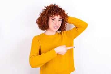 young redhead girl wearing yellow sweater over white background feeling positive has amazed expression, indicates something. One hand on head and pointing with other hand.
