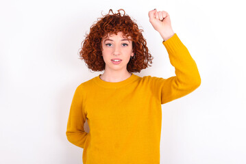 young redhead girl wearing yellow sweater over white background feeling serious, strong and rebellious, raising fist up, protesting or fighting for revolution.