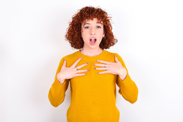 young redhead girl wearing yellow sweater over white background keeps hands on chest feeling shocked and scared, mouth widely opened, stares at camera saying: Who, me?