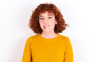 young redhead girl wearing yellow sweater over white background being nervous and scared biting lips looking camera with impatient expression, pensive.