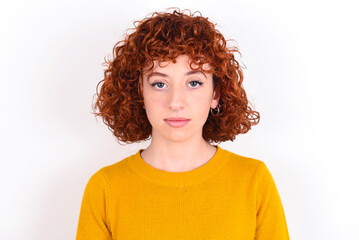 Joyful young redhead girl wearing yellow sweater over white background looking to the camera, thinking about something. Both arms down, neutral facial expression.