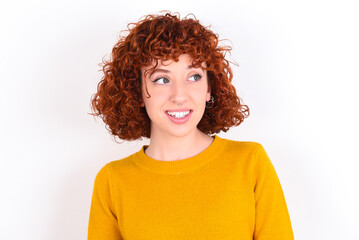 Oops! Portrait of young redhead girl wearing yellow sweater over white background  clenches teeth and looks confusedly aside, realizes her bad mistake,