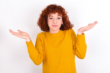 Puzzled and clueless young redhead girl wearing yellow sweater over white background with arms out, shrugging shoulders, saying: who cares, so what, I don't know.