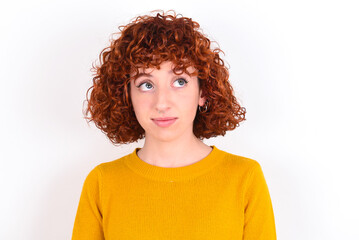 young redhead girl wearing yellow sweater over white background has worried face looking up lips together, being upset thinking about something important, keeps hands down.