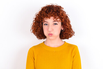 young redhead girl wearing yellow sweater over white background puffing cheeks with funny face. Mouth inflated with air, crazy expression.