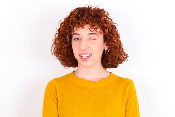 young redhead girl wearing yellow sweater over white background blinking eyes with pleasure having happy expression. Facial expressions and people emotions concept.