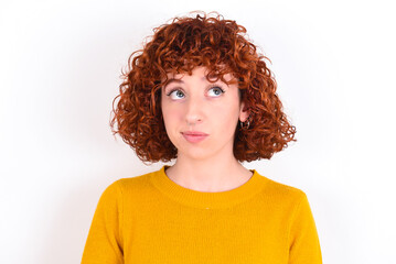young redhead girl wearing yellow sweater over white background crosses eyes, puts lips, makes grimace with awkward expression has fun alone, plays fool.