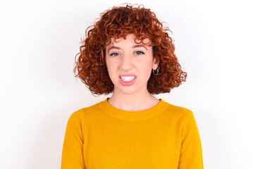 Mad crazy young redhead girl wearing yellow sweater over white background clenches teeth angrily, being annoyed with coming noise. Negative feeling concept.