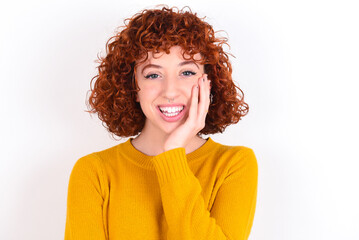 Shocked, astonished young redhead girl wearing yellow sweater over white back looking surprised in full disbelief wide open mouth with hand near face. Positive emotion facial expression body language.