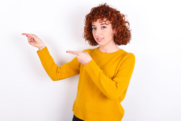young redhead girl wearing yellow sweater over white background points aside with  surprised expression with mouth opened, shows something amazing. Advertisement concept.